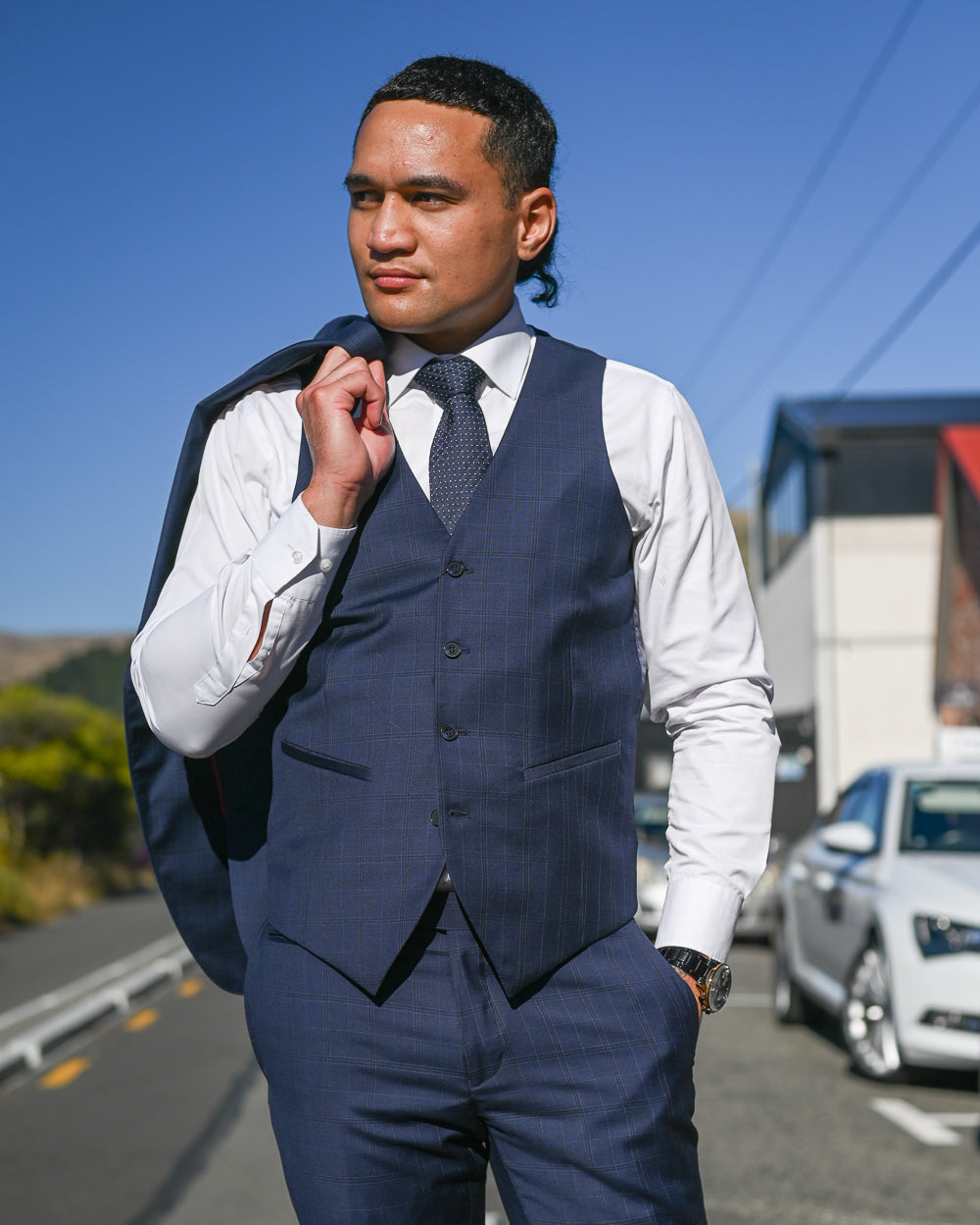 Handsome young Samoan man wearing a navy waistcoat with navy suit trousers and a navy jacket by Savile Row slung over his shoulder